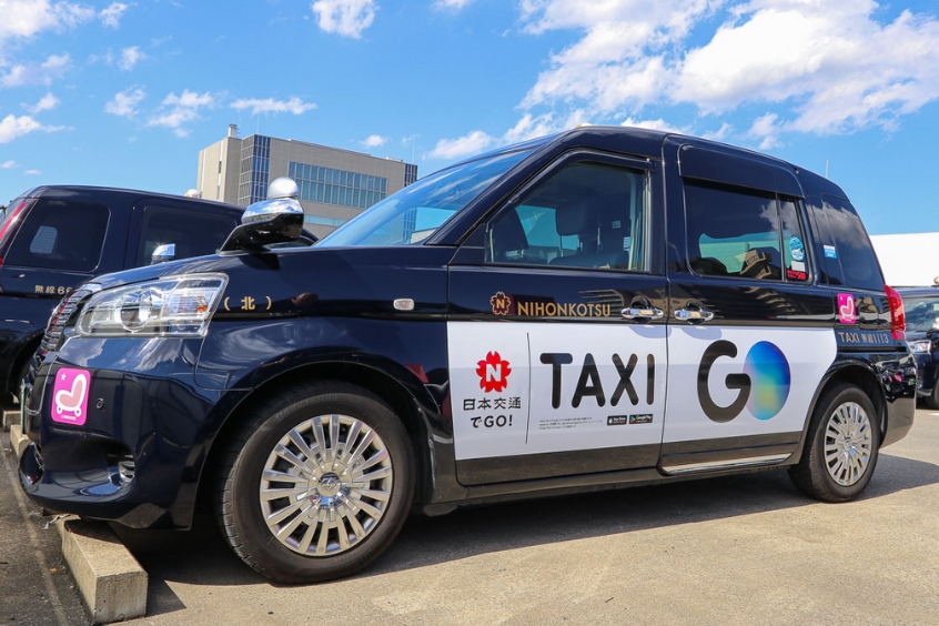 Taxi Nhật Bản với biểu tượng "TAXI GO" di chuyển trên đường phố Matsuyama.