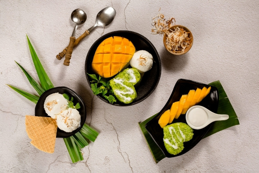 Món tráng miệng Thái Lan hấp dẫn với xoài tươi, kem dừa, bánh mochi xanh và bánh waffle, trang trí lá dứa trên bàn ăn phong cách nhiệt đới